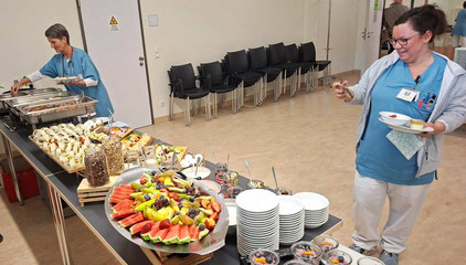 Zwei Pflegerinnen in blauer Funktionskleidung stehen in einem Raum mit einem mit warmen und kalten Speisen, Obst, Müsli und Gebäck gedeckten Buffet am Internationalen Tag der Pflegenden. Zwei Pflegerinnen in blauer Funktionskleidung stehen in einem Raum mit einem mit warmen und kalten Speisen, Obst, Müsli und Gebäck gedeckten Buffet am Internationalen Tag der Pflegenden.