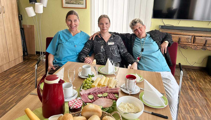 Drei Pflegekräfte in Funktionskleidung sitzen nebeneinander auf einem Sofa an einem gedeckten Frühstückstisch und blicken in die Kamera beim Internationalen Tag der Pflegenden im Immanuel Haus am Kalksee in Rüdersdorf bei Berlin. Drei Pflegekräfte in Funktionskleidung sitzen nebeneinander auf einem Sofa an einem gedeckten Frühstückstisch und blicken in die Kamera beim Internationalen Tag der Pflegenden im Immanuel Haus am Kalksee in Rüdersdorf bei Berlin.